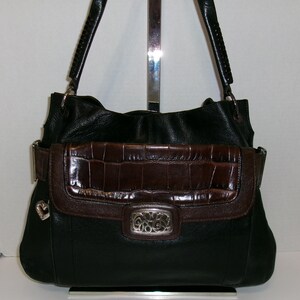 K&ouml;nnte beinhalten: Schwarze Lederhandtasche mit braunem Kunstkrokodil-Klappverschluss und silberfarbenen Beschl&auml;gen. Die Tasche hat geflochtene Griffe und einen dekorativen silbernen Herzanh&auml;nger. Der Klappverschluss hat einen silbernen Verschluss mit einem verzierten Design.