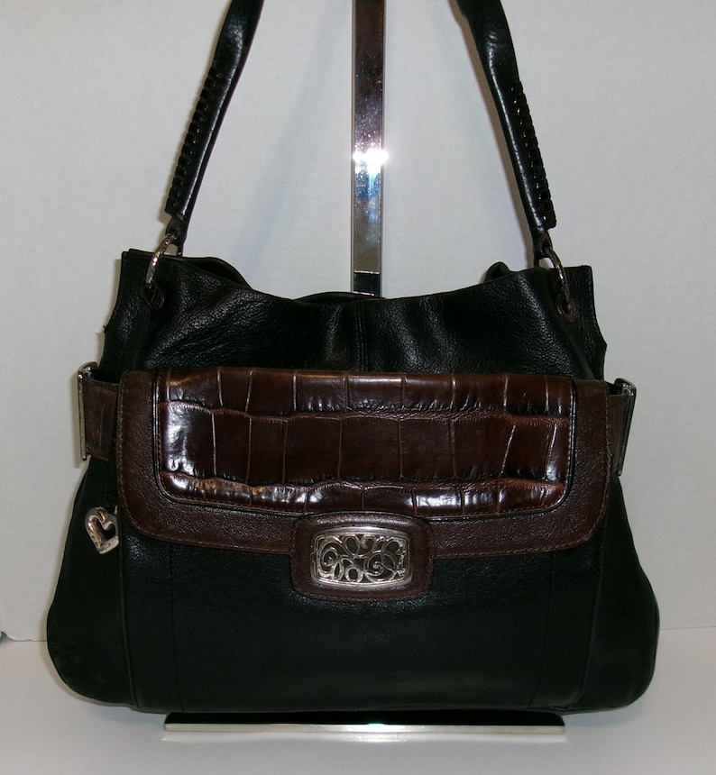 K&ouml;nnte beinhalten: Schwarze Lederhandtasche mit braunem Kunstkrokodil-Klappverschluss und silberfarbenen Beschl&auml;gen. Die Tasche hat geflochtene Griffe und einen dekorativen silbernen Herzanh&auml;nger. Der Klappverschluss hat einen silbernen Verschluss mit einem verzierten Design.