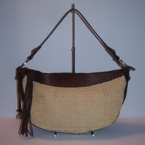 Może przedstawiać: Brązowa sk&oacute;rzana torebka ze słomy z długim brązowym sk&oacute;rzanym paskiem i frędzlami. Torebka znajduje się na stojaku.