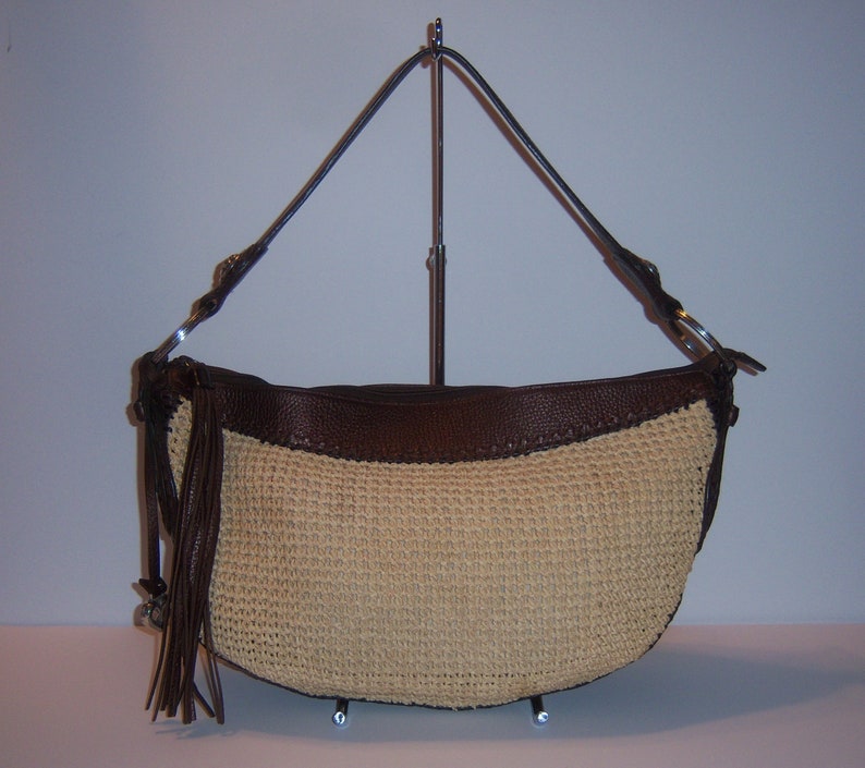 Może przedstawiać: Brązowa sk&oacute;rzana torebka ze słomy z długim brązowym sk&oacute;rzanym paskiem i frędzlami. Torebka znajduje się na stojaku.