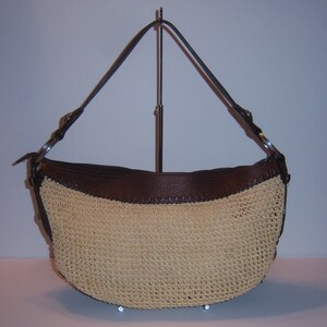 Może przedstawiać: Brązowa sk&oacute;rzana torebka ze słomianym korpusem i brązowymi sk&oacute;rzanymi wykończeniami. Torebka ma jeden pasek i jest prezentowana na stojaku.