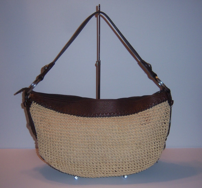 Może przedstawiać: Brązowa sk&oacute;rzana torebka ze słomianym korpusem i brązowymi sk&oacute;rzanymi wykończeniami. Torebka ma jeden pasek i jest prezentowana na stojaku.