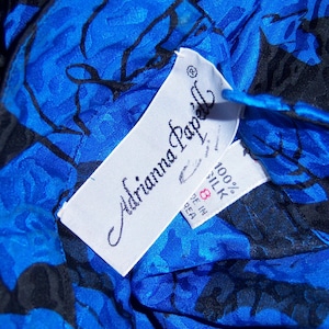 May include: Close-up of a blue silk garment with a black floral pattern. The garment has a tag that reads "Adrianna Papell" and "100% Silk". The tag also indicates the size is 8.