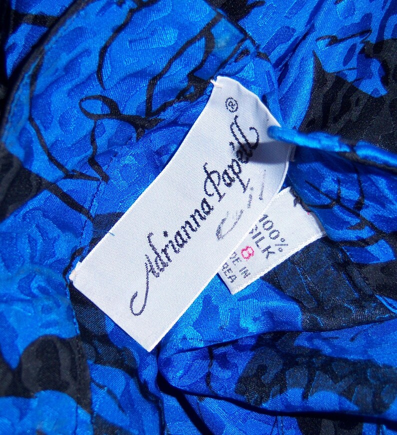 May include: Close-up of a blue silk garment with a black floral pattern. The garment has a tag that reads "Adrianna Papell" and "100% Silk". The tag also indicates the size is 8.