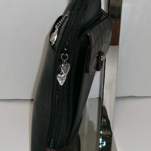 May include: Black leather handbag with a zip closure and a small, textured brown pocket. The bag features silver heart-shaped charms and a matching brown base. The bag is displayed on a silver stand.