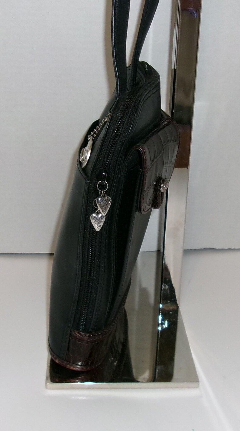 May include: Black leather handbag with a zip closure and a small, textured brown pocket. The bag features silver heart-shaped charms and a matching brown base. The bag is displayed on a silver stand.
