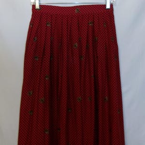 May include: A red skirt with a black grid pattern and small, scattered floral designs. The skirt is hanging on a metal hanger against a white background. The skirt has pleats and a high waist.