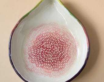 May include: A teardrop-shaped ceramic dish with a white interior featuring a central pink radial pattern. The outer rim is painted with a gradient of green, purple, and red tones, creating a colorful border around the smooth, concave surface.