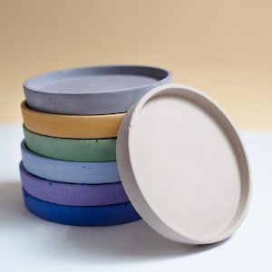 May include: A stack of seven round, concrete trays in various colors: light gray, yellow, green, blue, purple, and navy. One off-white tray leans against the stack. The trays are on a white surface with a beige background.