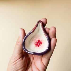 May include: A small, fig-shaped ceramic dish with a dark purple outer rim and a cream-colored interior. The center of the dish features a vibrant red circular pattern with small white dots, mimicking the appearance of the inside of a sliced fig.