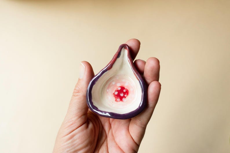 May include: A small, fig-shaped ceramic dish with a dark purple outer rim and a cream-colored interior. The center of the dish features a vibrant red circular pattern with small white dots, mimicking the appearance of the inside of a sliced fig.