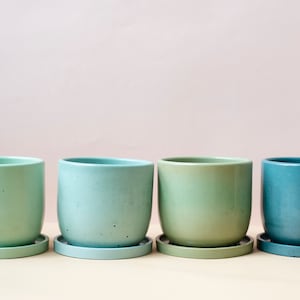 May include: Four ceramic plant pots in shades of green and blue, each with a matching saucer. The pots are cylindrical with a smooth, matte finish. The colors range from light green to teal, creating a gradient effect. Ideal for indoor plants.