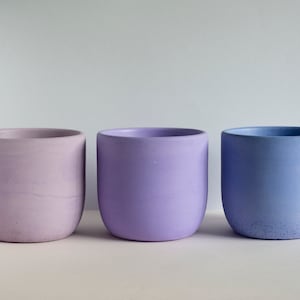 May include: Three pastel-colored, cylindrical planters. The planters are in shades of lavender, purple, and blue. The planters have a smooth, matte finish and a rounded base. They are arranged in a row against a light gray background.