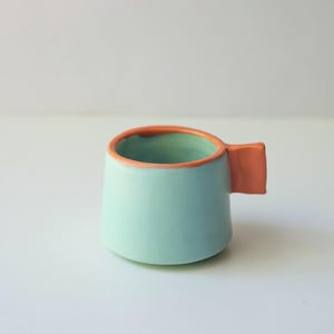 May include: A small ceramic mug with a smooth, light mint green exterior and interior. The rim of the mug is painted in a vibrant coral orange, which matches the rectangular, flat-sided handle attached to the side of the vessel.