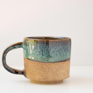 May include: A ceramic mug featuring a two-tone design with a speckled tan base and a glossy, mottled teal and dark brown upper section. The mug has a sturdy, curved handle and a slightly recessed base, showcasing a reactive glaze finish.