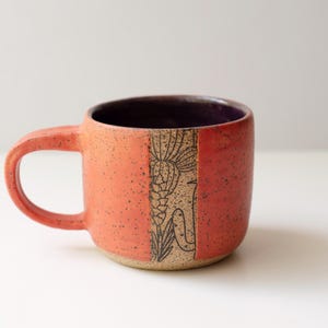 May include: A ceramic mug with a speckled orange exterior and a dark purple interior. The mug features a vertical band with a black line drawing of a cactus and desert flora. It has a rounded handle on the left side and a smooth, curved body.