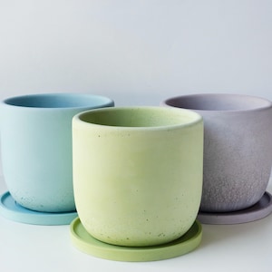 May include: Three pastel-colored ceramic plant pots with matching saucers. The pots are cylindrical with rounded bottoms. Colors include light blue, green, and lavender. The pots are sitting on a white surface.