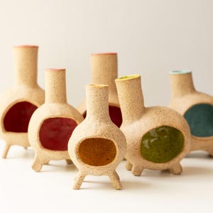 May include: A collection of five small, speckled ceramic vases with tripod legs and tall necks. Each vase features a hollow, rounded body with a smooth, brightly colored interior glaze in shades of red, yellow, green, and blue.