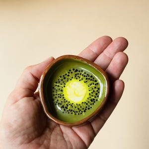 May include: A small, hand-formed ceramic dish with an irregular, rounded shape. The interior features a painted kiwi fruit design with a bright yellow center, a light green ring, and small black dots radiating outward. The outer rim is brown.