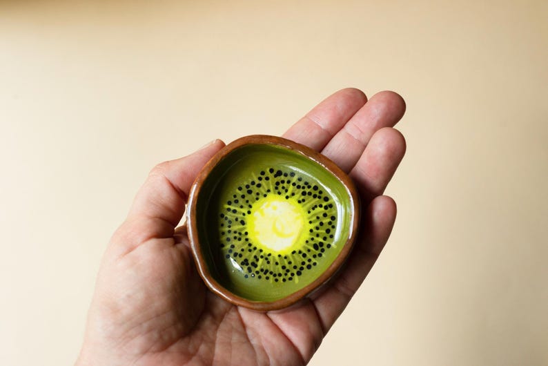 May include: A small, hand-formed ceramic dish with an irregular, rounded shape. The interior features a painted kiwi fruit design with a bright yellow center, a light green ring, and small black dots radiating outward. The outer rim is brown.