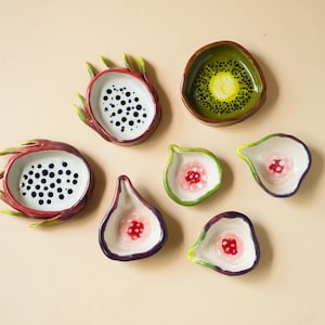 May include: A collection of seven ceramic dishes shaped like fruit slices. Two are dragon fruit bowls with white interiors and black dots, three are fig-shaped dishes with pink centers, one is a kiwi slice bowl, and one is a small white dish.