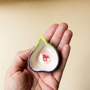 May include: A small, fig-shaped ceramic dish with a smooth, cream-colored interior featuring a central red and white circular pattern. The exterior rim is painted with a vibrant green and deep purple gradient, creating a distinct fruit-like appearance.