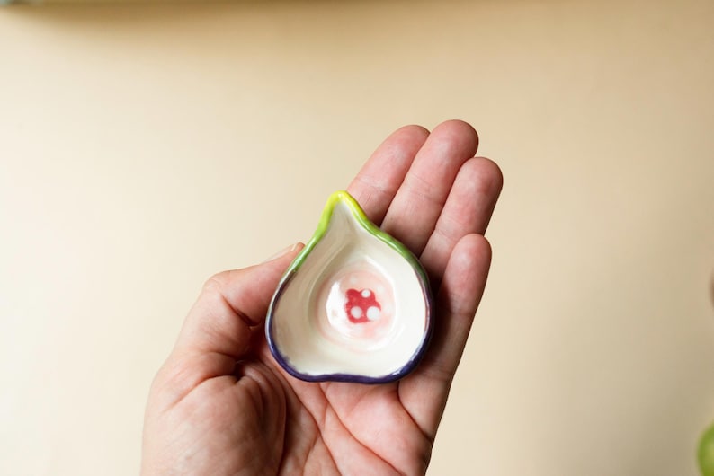 May include: A small, fig-shaped ceramic dish with a smooth, cream-colored interior featuring a central red and white circular pattern. The exterior rim is painted with a vibrant green and deep purple gradient, creating a distinct fruit-like appearance.
