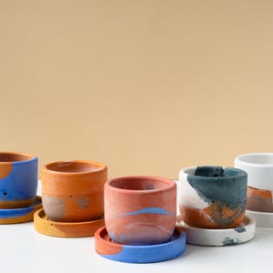 May include: Five small cylindrical planters with matching saucers, each featuring a unique marbled or color-blocked design in shades of blue, orange, terracotta, white, and dark gray. The pots have a smooth, matte finish and a simple, open top.
