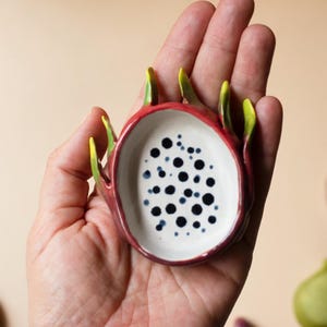 May include: A small, oval-shaped ceramic dish designed to resemble a sliced dragon fruit. The exterior is a deep magenta-red with pointed green-tipped scales, while the interior is white with numerous small, dark blue-black circular spots.