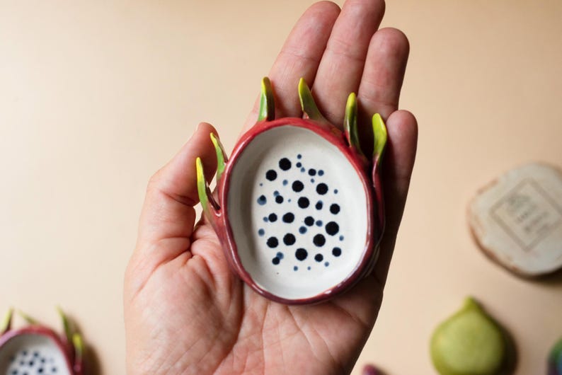 May include: A small, oval-shaped ceramic dish designed to resemble a sliced dragon fruit. The exterior is a deep magenta-red with pointed green-tipped scales, while the interior is white with numerous small, dark blue-black circular spots.