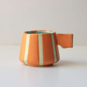 May include: A ceramic mug featuring a tapered, wide-bottomed shape with vertical stripes in alternating shades of bright orange and pale mint green. The mug includes a single, flat, rectangular handle extending from the side of the vessel.