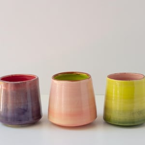 May include: Three ceramic tumblers with a smooth, tapered silhouette. The left tumbler features a gradient of deep purple and magenta. The center tumbler is a soft pastel pink with a lime green interior. The right tumbler is a bright yellow-green.
