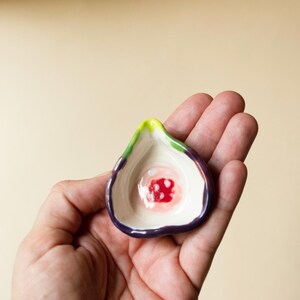 May include: A small, hand-held ceramic dish shaped like a halved fig. The exterior features a deep purple skin, while the interior is cream-colored with a vibrant red and white center, accented by a bright lime green stem at the pointed top edge.