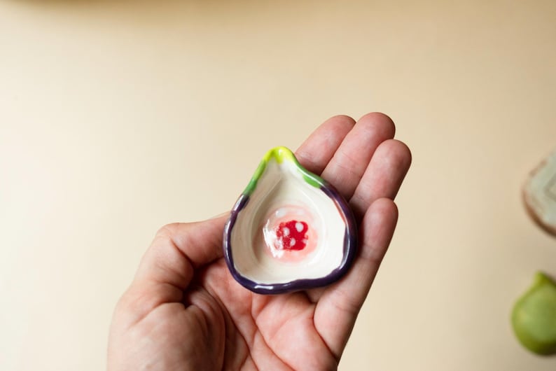 May include: A small, hand-held ceramic dish shaped like a halved fig. The exterior features a deep purple skin, while the interior is cream-colored with a vibrant red and white center, accented by a bright lime green stem at the pointed top edge.