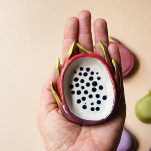 May include: A small ceramic dish shaped like a halved dragon fruit with a white interior featuring black painted dots and a vibrant pink and green exterior. The dish has curved, pointed edges mimicking the skin of the fruit.