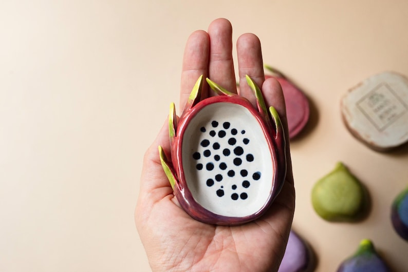 May include: A small ceramic dish shaped like a halved dragon fruit with a white interior featuring black painted dots and a vibrant pink and green exterior. The dish has curved, pointed edges mimicking the skin of the fruit.