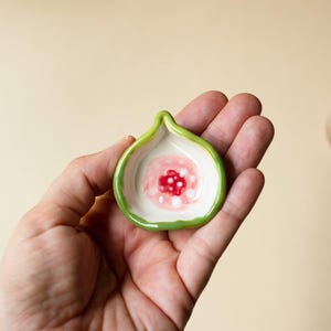May include: A small, fig-shaped ceramic dish with a glossy green rim and a white interior. The center of the dish features a painted design of a red circular core surrounded by soft pink and white dots, mimicking the appearance of a sliced fig.
