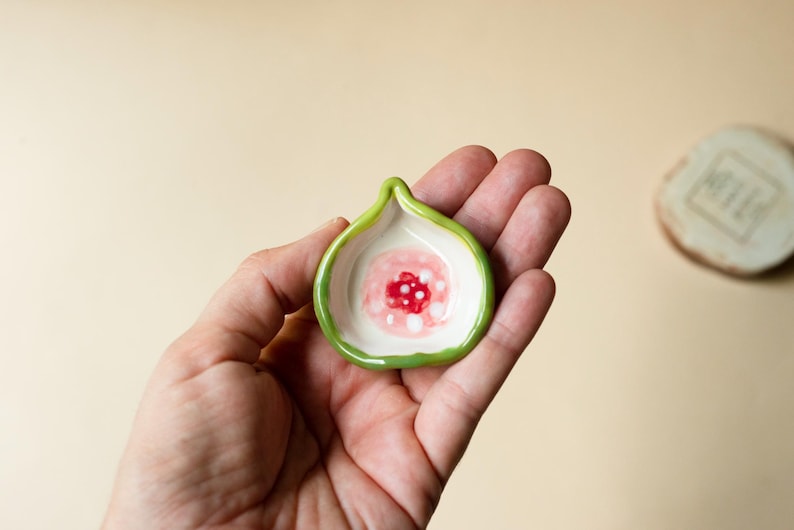 May include: A small, fig-shaped ceramic dish with a glossy green rim and a white interior. The center of the dish features a painted design of a red circular core surrounded by soft pink and white dots, mimicking the appearance of a sliced fig.