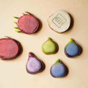 May include: Two circular dragon fruit slices with green-tipped edges, four teardrop-shaped fig pieces in shades of green and purple, and a round beige ceramic disc stamped with the text COOL HAND ROBYN on a light tan background.