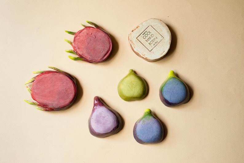 May include: Two circular dragon fruit slices with green-tipped edges, four teardrop-shaped fig pieces in shades of green and purple, and a round beige ceramic disc stamped with the text COOL HAND ROBYN on a light tan background.