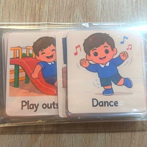 Puede incluir: Un juego de tarjetas didácticas con ilustraciones de dibujos animados de niños. Una tarjeta muestra a un niño en un tobogán, la otra muestra a un niño bailando con notas musicales. Las palabras "Play outs" y "Dance" están impresas en las tarjetas.