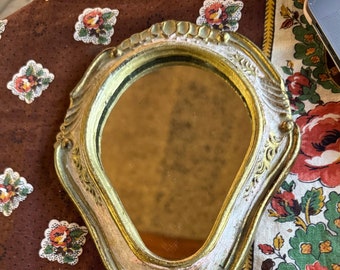 Puede incluir: Un espejo de mano vintage con un marco dorado y blanco adornado. El espejo tiene una forma ligeramente ovalada y un mango largo.
