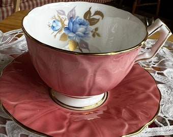 Può includere: Un set da tè rosa con un design floreale che presenta una rosa blu. Il set ha un bordo dorato.