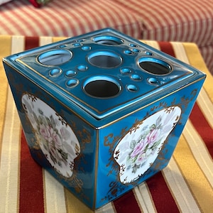 Puede incluir: Un jarrón de cerámica cuadrado con un esmalte azul turquesa y ribetes dorados. El jarrón tiene seis agujeros para tallos de flores y un diseño floral en cada lado.