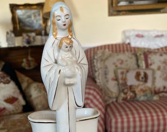 Op de afbeelding: Een wit keramiek beeldje van de Maagd Maria die een baby Jezus vasthoudt. Het beeldje is versierd met gouden accenten en blauwe details. Het beeldje staat op een zwarte voet.