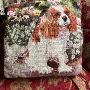 Puede incluir: Una almohada de punto de aguja marr&oacute;n, blanca y tostada con un perro Cavalier King Charles Spaniel rodeado de flores. La almohada est&aacute; sobre una silla con estampado rojo y dorado.