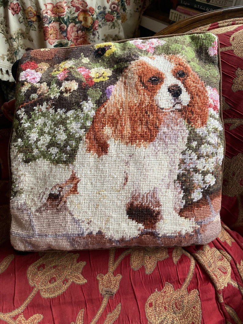 Puede incluir: Una almohada de punto de aguja marr&oacute;n, blanca y tostada con un perro Cavalier King Charles Spaniel rodeado de flores. La almohada est&aacute; sobre una silla con estampado rojo y dorado.