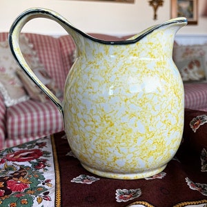 Puede incluir: Una jarra de cerámica amarilla y blanca moteada con un borde y un asa negros. La jarra está sentada sobre un mantel con estampado floral.