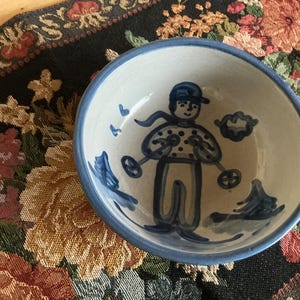 May include: A round ceramic bowl with a cream-colored base and a blue rim. The interior features a hand-painted blue illustration of a person wearing a cap and polka-dot shirt, holding ski poles, surrounded by stylized clouds and trees.