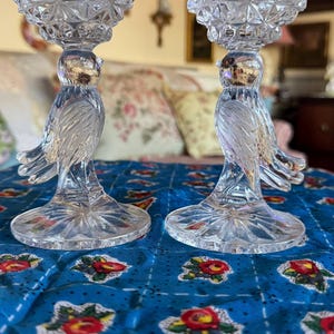 May include: Two crystal candle holders shaped like birds with a clear glass base and a geometric pattern on the top. The candle holders are sitting on a blue floral tablecloth.